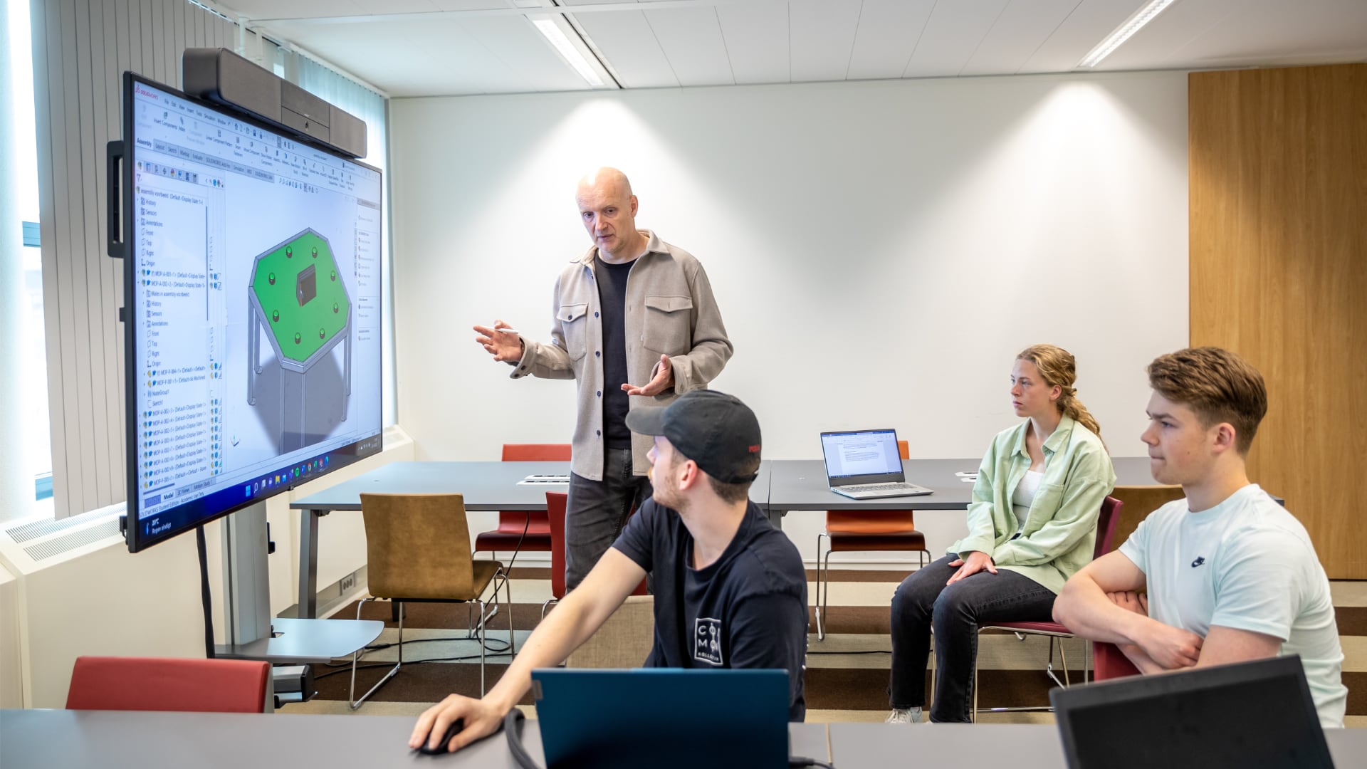 Docent geeft uitleg aan 3 studenten met een uitwerking op een groot scherm